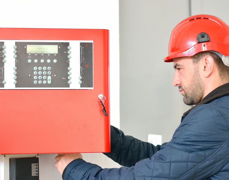 A man in red hard hat holding up a fire alarm.