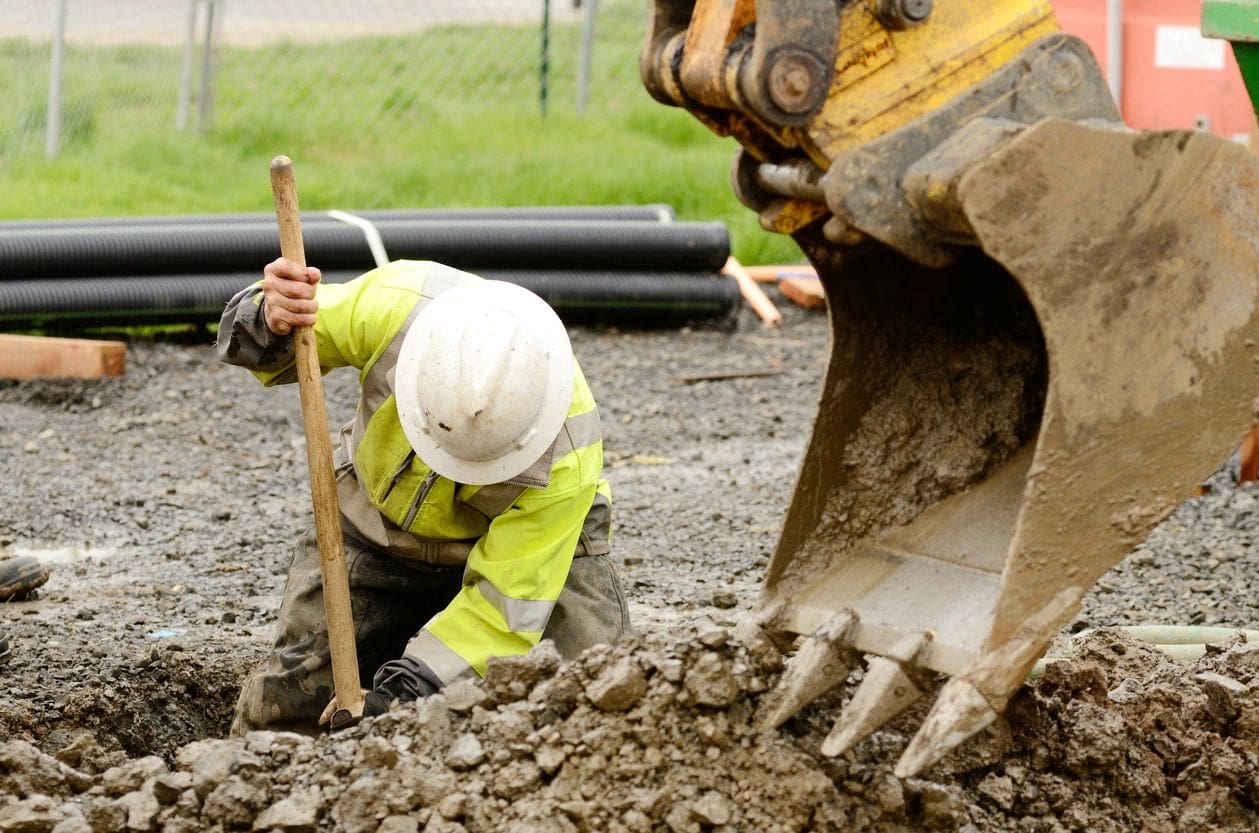 A man in yellow jacket digging dirt with shovel.